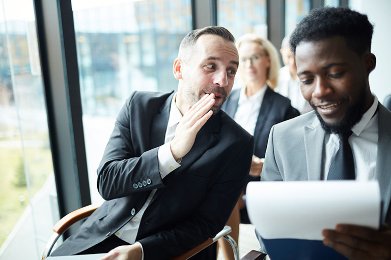 Two men whispering to eachother while one holds a paper.